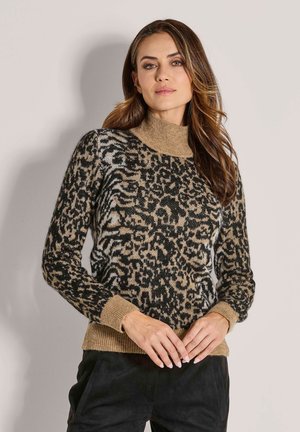 Frau mit langen, welligen Haaren, die einen beigen und schwarzen Leopardenmuster-Pullover mit Stehkragen und schwarze Hosen trägt, vor einem schlichten Hintergrund stehend.