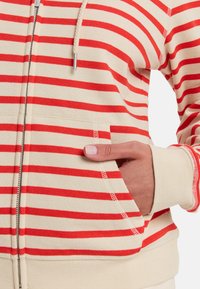 Sudadera con cremallera con rayas horizontales en crema y rojo, que incluye un bolsillo frontal y puños acanalados. Tela suave con un corte relajado.