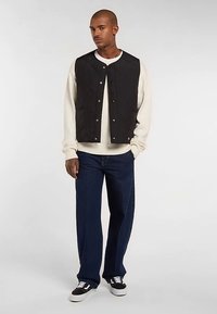 Veste matelassée noire à boutons-pression, portée sur un sweat crème, associée à un jean bleu foncé et des baskets noires. Design simple et fonctionnel.