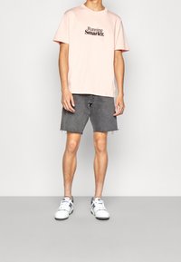 Camiseta de algodón de color rosa claro con "Running Smarter" impreso en negro, combinada con pantalones cortos de mezclilla gris. Zapatillas blancas con detalles grises.