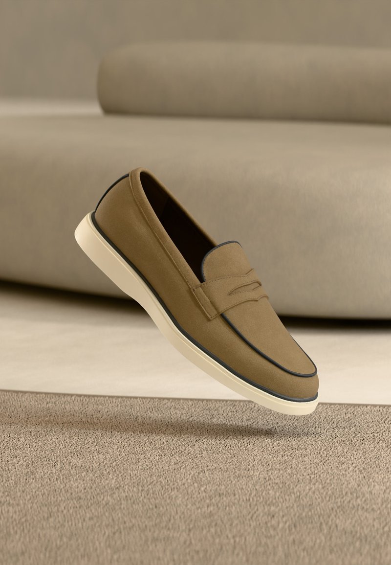 Mocassin en daim marron clair avec semelle blanche flottant au-dessus d'un tapis beige, canapé aux tons neutres en arrière-plan flou.