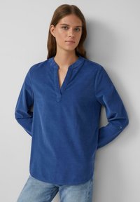 Blouse en coton bleu avec col en V, manches longues et détails de boutons aux poignets. Tissu texturé et design d'ourlet arrondi.
