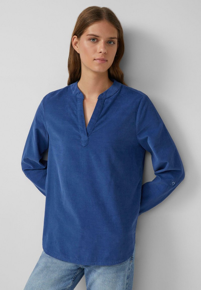 Blouse en coton bleu avec col en V, manches longues et détails de boutons aux poignets. Tissu texturé et design d'ourlet arrondi.
