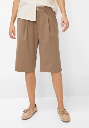 Donna con pantaloncini marroni plissettati fino al ginocchio, mocassini beige con lacci e top bianco parzialmente visibile, in piedi contro uno sfondo bianco.