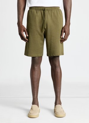 Uomo che indossa pantaloncini verde oliva con coulisse e scarpe beige slip-on in piedi su sfondo bianco.