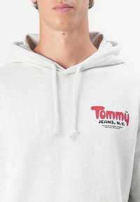 Lys grå sweatshirt lavet af bomuld, med et rødt "Tommy Jeans" logo på brystet. Inkluderer en snor og en kængurulomme.