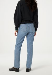 Persoon met krullend haar, gekleed in een zwarte sweater, lichtblauwe spijkerbroek en zwarte schoenen met studs, staand met de rug naar de camera op een effen achtergrond.