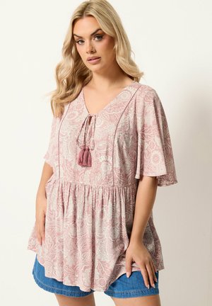 Kvinde med langt blondt hår iført en løs pink og hvid paisley-mønstret bluse med kvastbind og denimshorts, stående mod en hvid baggrund.