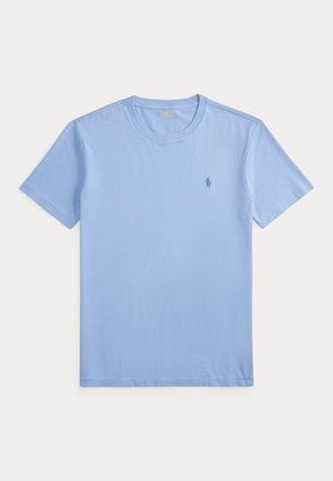 Jeune homme aux cheveux bouclés, vêtu d'un t-shirt bleu, d'un jean foncé, de chaussures marron et d'une montre-bracelet, debout devant un fond neutre uni.