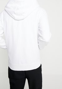 Person som bär en enkel vit hoodie med huvan nere och svarta byxor, visad bakifrån mot en vit bakgrund.