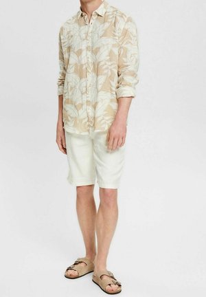 Uomo in piedi che indossa una camicia a maniche lunghe beige con motivo floreale, pantaloncini bianchi fino al ginocchio e sandali beige con fibbia su sfondo bianco.