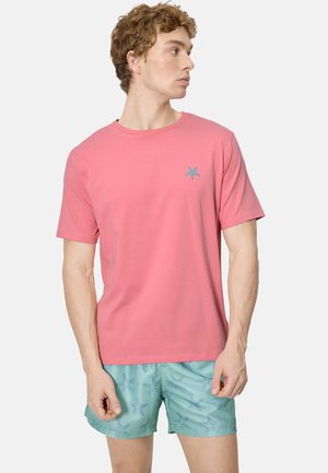 Hombre con una camisa rosa de manga corta con un pequeño logo de estrella de mar y pantalones cortos azul claro con un patrón de estrellas de mar, mirando hacia la derecha.