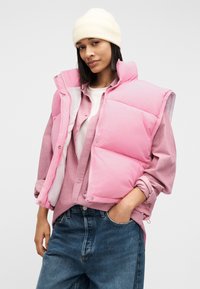 Γυναίκα που φοράει λευκό σκουφάκι, ροζ μπουφάν τύπου puffer, ροζ πουκάμισο, λευκό μπλουζάκι και μπλε τζιν, με το ένα χέρι στην τσέπη, μπροστά σε απλό φόντο.