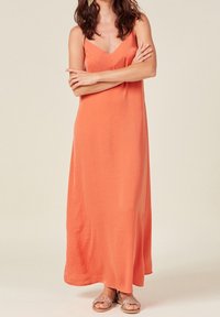 Maxikleid in Korallenorange, aus leichtem, glattem Stoff. Mit V-Ausschnitt und verstellbaren Spaghetti-Trägern. Getragen mit hellbraunen Sandalen.