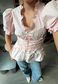 Femme portant un chemisier rose clair à volants avec des manches bouffantes, un jean bleu et des ballerines blanches, debout à l'intérieur.