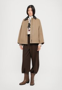 Veste extérieure marron avec un col noir, associée à un pull à motifs blancs, un pantalon large marron et des bottes en cuir marron.