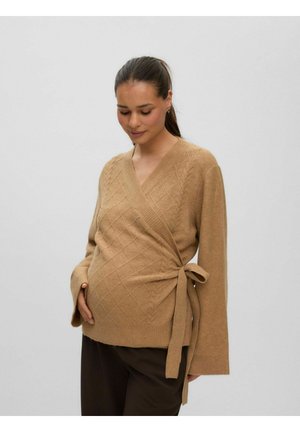 Femme enceinte portant un pull cache-cœur camel avec un motif en losanges et des manches larges, debout devant un fond clair uni.