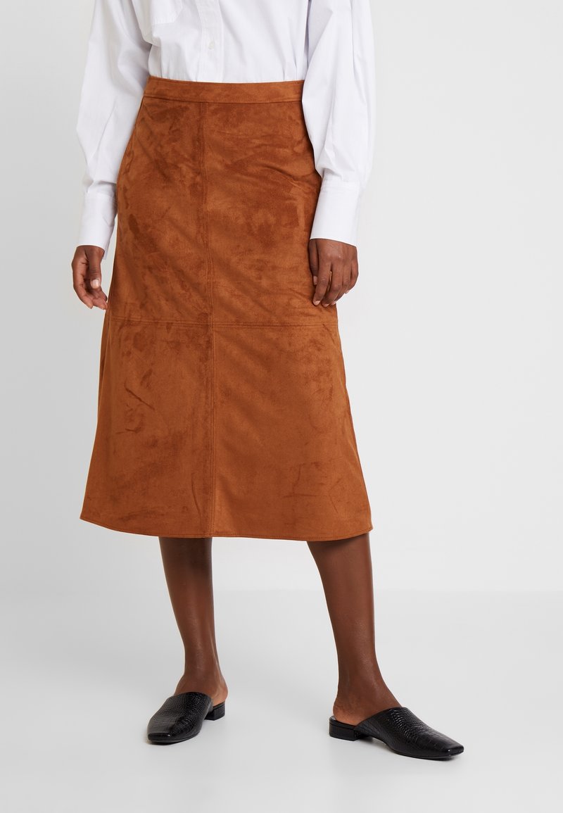 Femme portant une jupe en daim marron longueur genoux, un chemisier blanc à manches longues et des chaussures plates noires sans lacets, debout sur un sol gris.