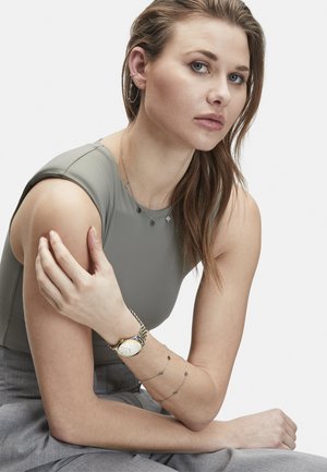 Silberner Schmuck, geschichtet mit kleinen geometrischen Charms, graues eng anliegendes Oberteil, aufwändige Uhr mit Gold- und Silberelementen, graue Hose.