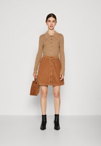 Bruine geribbelde lange mouw top met kraag, gecombineerd met een tan corduroy rok met knopen aan de voorkant, en zwarte enkellaarsjes.