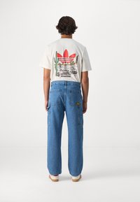 Camiseta corta de manga corta color crema con el logo rojo de Adidas y un gráfico del desierto. Pantalones cargo de mezclilla azul con múltiples bolsillos y un corte relajado. Zapatos blancos.