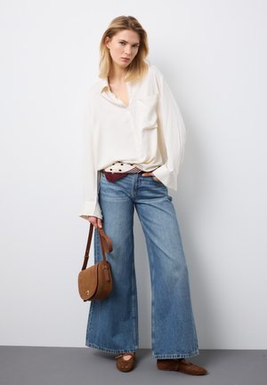 Mujer con blusa blanca holgada, vaqueros azules de pierna ancha, zapatos planos marrones tejidos, sosteniendo un bolso de hombro de ante marrón contra una pared blanca.