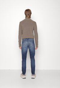 Person mit hellbraunen Haaren, die einen taupefarbenen Langarmpullover, blaue Jeans und beige Sneakers trägt, und vor einer weißen Wand steht.