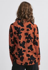 Blusa color ruggine con un motivo a foglia nera, maniche lunghe e una superficie texturizzata. L'orlo è arrotondato con spacchi laterali.