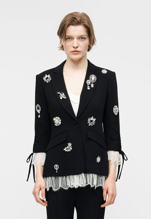 Γυναίκα με μαύρο σακάκι διακοσμημένο με διάφορα ασημένια και μαργαριταρένια κονκάρδες, λευκό δαντελένιο πουκάμισο ορατό στα μανίκια και στο τελείωμα.