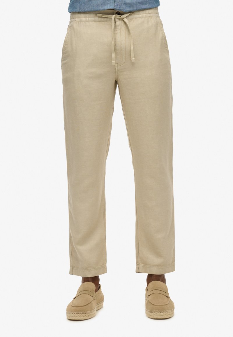 Pantalon en lin beige avec une coupe décontractée, une taille élastique et un cordon de serrage. Caractérisé par des poches latérales et des poignets fuselés, associé à des mocassins beiges.