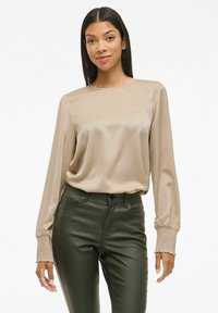 Beige satinbluse med lange ermer, med ribbede mansjetter og en løs passform, kombinert med mørkegrønne tettsittende bukser. Glatt tekstur.