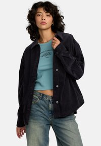 Zwarte corduroy overhemdjas met zilveren hardware, met een losse pasvorm. Gedragen over een blauwe geribde crop top en gecombineerd met licht gewassen jeans.