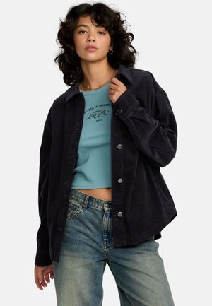 Camicetta giacca in corduroy nera con hardware argentato, caratterizzata da una vestibilità rilassata. Indossata sopra un top corto a costine blu e abbinata a jeans chiari.