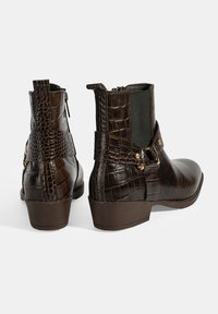 Bottines marron avec un design gaufré crocodile, fermeture zippée, panneaux latéraux élastiques et accents en métal doré sur la sangle. Talon plat.