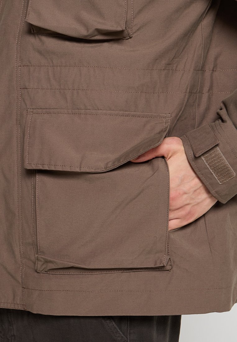 Giacca marrone resistente all'acqua con grande tasca frontale, dettaglio con velcro ai polsi e texture liscia. Design funzionale e pratico.