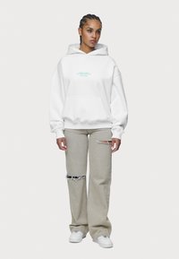 Bílá mikina s kapucí a klokaní kapsou, se tyrkysovým textem. Vytvořeno s světle šedými vybledlými džíny a bílými teniskami.