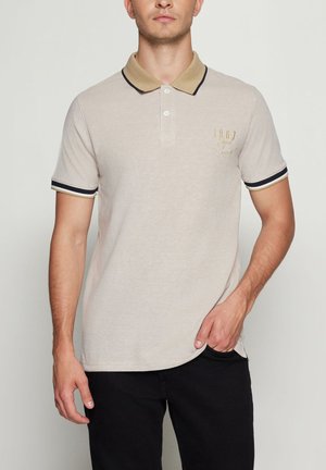 Mand iført beige polo med tan krave og sorte stribede ærmekanter, sorte bukser, hånd i lommen, mod ensfarvet baggrund.