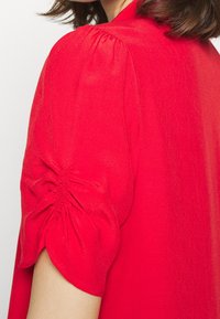 Blusa rossa con maniche a sbuffo e dettagli arricciati, realizzata in un tessuto leggero con una sottile texture.