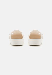 Hvide åbne sandaler med en glat, skinnende overflade, udstyret med en beige fodseng og justerbare stropper på siden. Flad sål design.