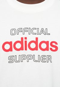 Bela majica s kratkimi rokavi s tiskom "URADNI dobavitelj adidas", beseda "adidas" je napisana z odebeljenim rdečim fontom, ostale besede pa z tankim črnim obrobnim fontom.