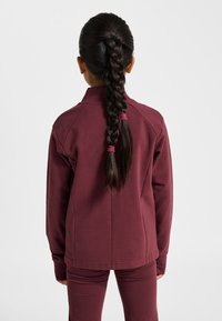 Vinröd jacka av mjukt tyg med ståkrage och raglanärmar, i kombination med matchande byxor. Bakifrån syns en fläta.