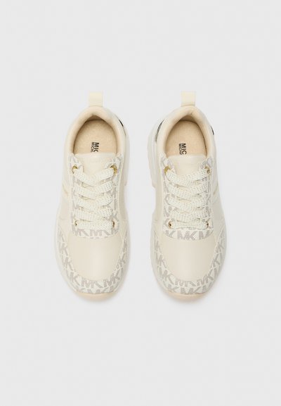 Zapatilla de color beige claro con detalles texturizados y una suela con patrón. Presenta cordones planos, herrajes en tono dorado y un collar acolchado para mayor comodidad.