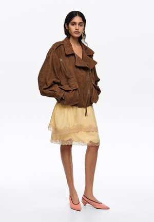 Mujer vistiendo una chaqueta grande de ante marrón, falda amarilla con encaje en el borde y zapatos rosas de tacón bajo, de pie contra un fondo blanco.