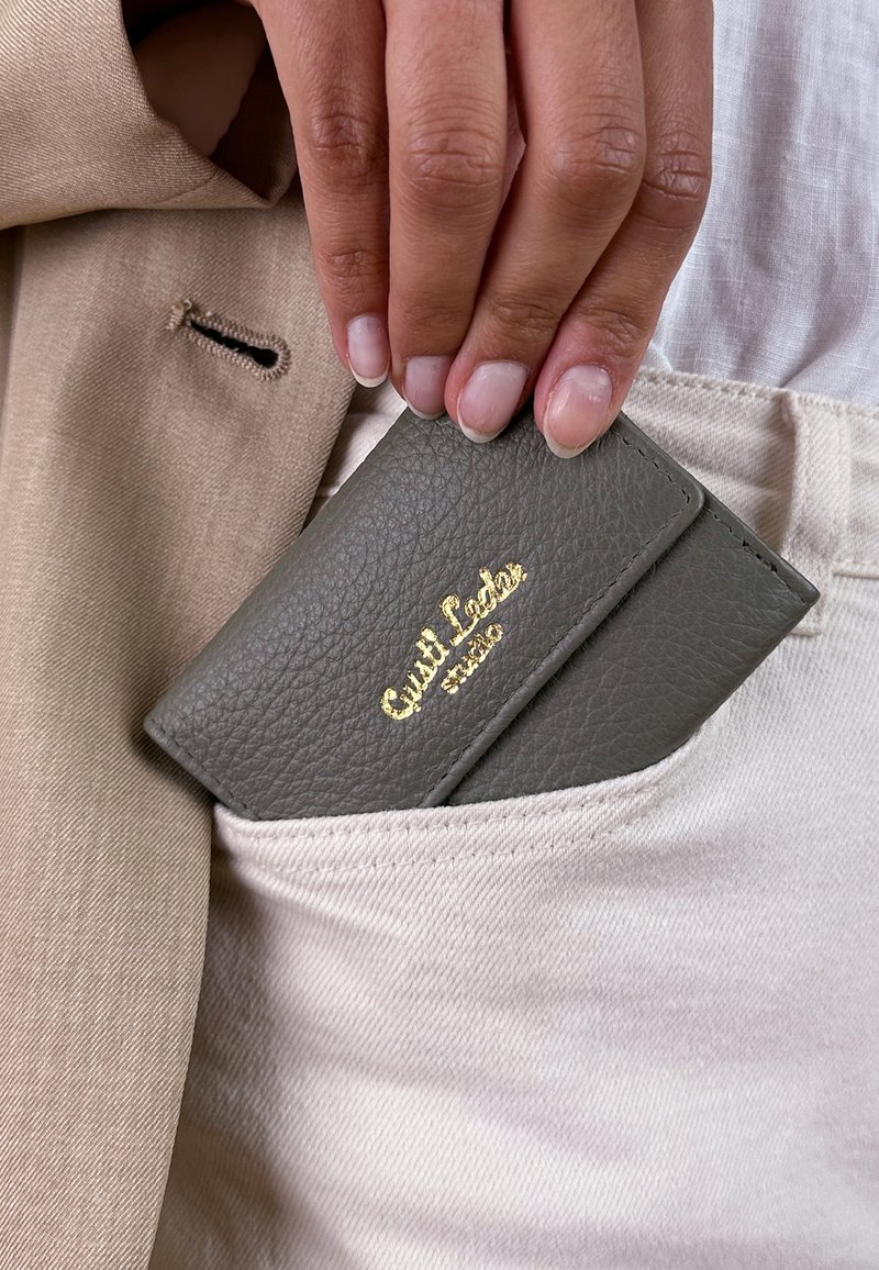 Hand mit natürlichen Nägeln, die eine graue Lederbrieftasche mit goldenen Buchstaben in die Tasche einer hellbeigen Hose steckt; ein ärmel einer beigen Jacke ist sichtbar.