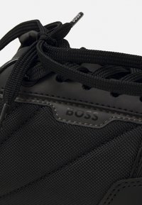 Gros plan d'une basket noire avec des lacets épais noués en nœud et une surface en tissu texturé montrant le nom de la marque "BOSS."