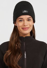 Sort strikket hue med ribbet tekstur og et logo-patch, båret med en lynlåset sort fleecejakke. Enkel design, ingen fremtrædende mønstre.
