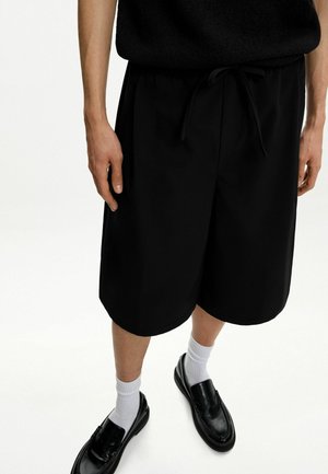 Schwarze, maßgeschneiderte Shorts aus glattem Stoff mit einem elastischen Bund und Kordelzug. Kombiniert mit weißen Socken und schwarzen Loafers.