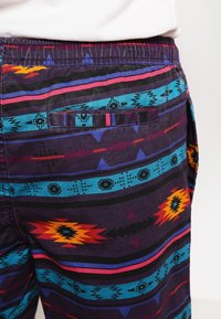 Pantalones cortos de patrón tribal multicolor con una base morada, que presentan detalles en azul, rojo y naranja. Incluyen un bolsillo trasero y una cintura elástica.