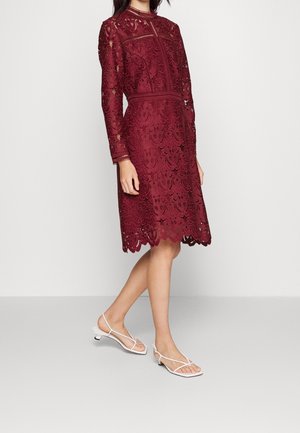 Abito di pizzo bordeaux con maniche lunghe, motivi floreali, orlo smerlato e vita aderente. Abbinato a sandali bianchi con cinturini.