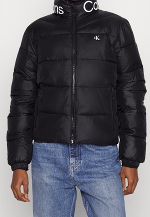 Mężczyzna w czarnej kurtce puchowej Calvin Klein z przednim zamkiem błyskawicznym i logo na klatce piersiowej oraz kołnierzu, w połączeniu z niebieskimi dżinsami.